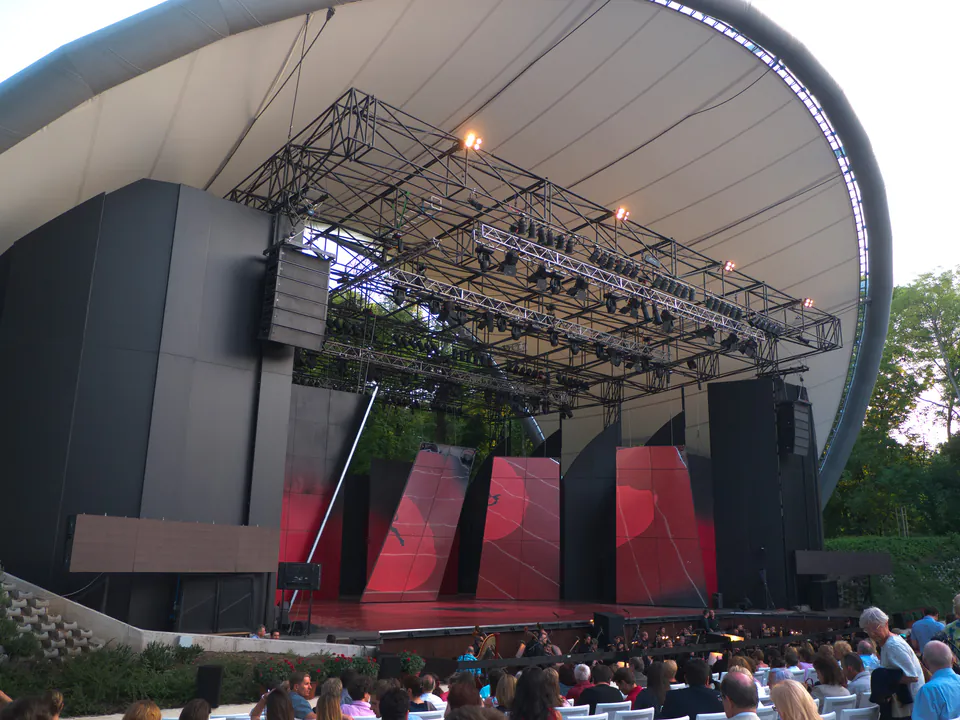 Modern outdoor amphitheater with curved roof, stage lighting, and audience seating during evening performance