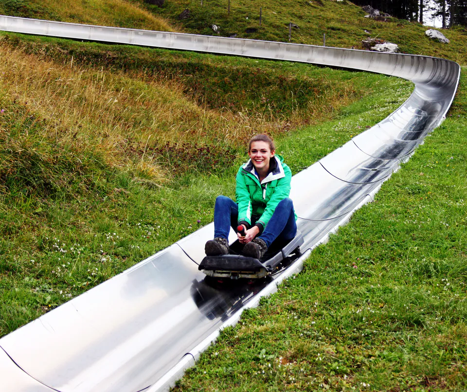 Curved metal toboggan track winding down grassy hillside with rider enjoying descent