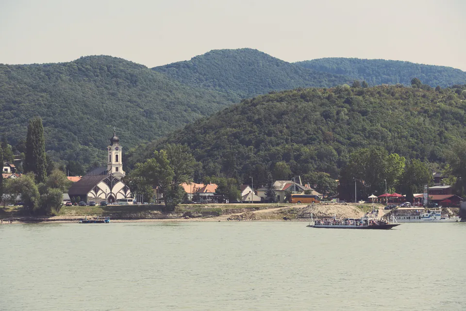 Ferry crossing Danube River with church spire and forested hills in background