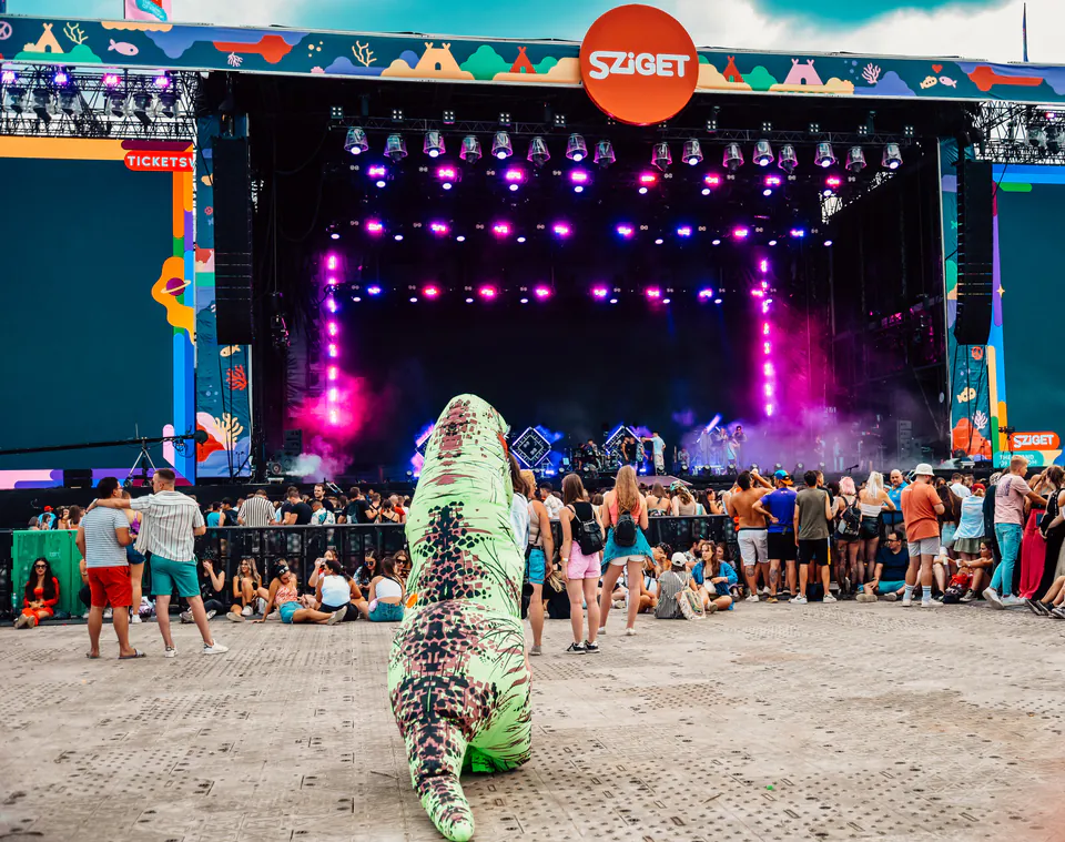 Festival stage with colorful lighting and crowd at Sziget Festival, with inflatable dinosaur costume in foreground