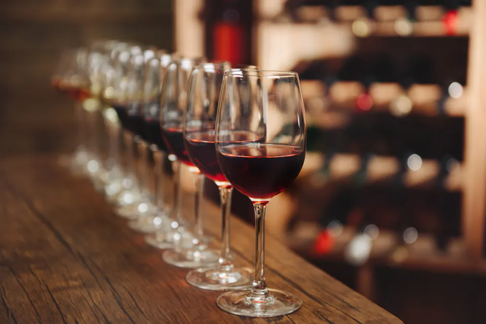 Line of wine glasses filled with red wine on wooden table with wine rack in background