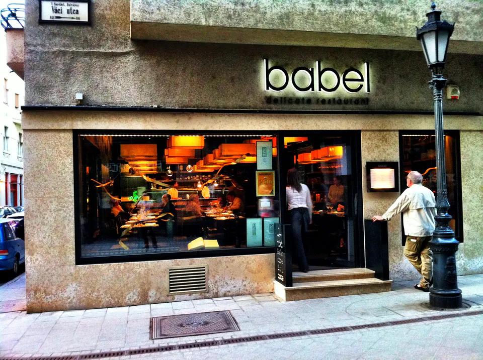 Exterior view of Babel restaurant with illuminated signage and large window showing dining room
