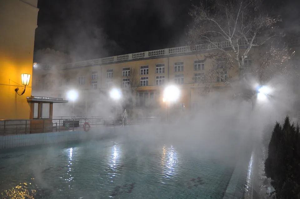 Steamy outdoor thermal pool at night with lights illuminating rising mist over water surface