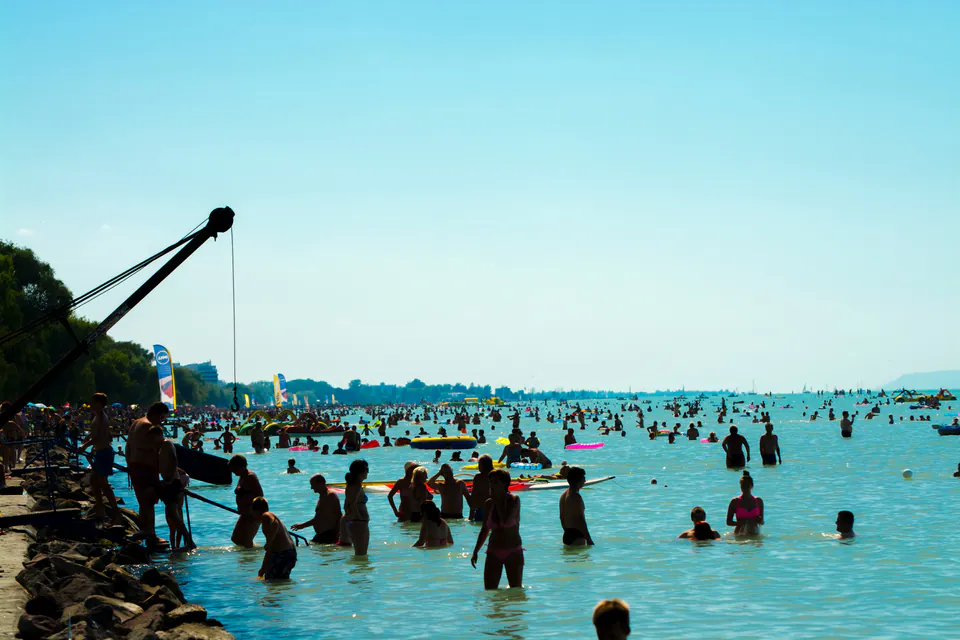 Crowded beach scene with people swimming in shallow waters and recreational facilities along shoreline