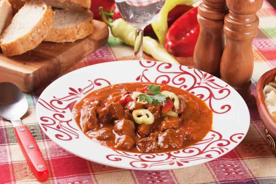 Traditional Hungarian goulash served in decorative plate with bread and peppers on checkered tablecloth