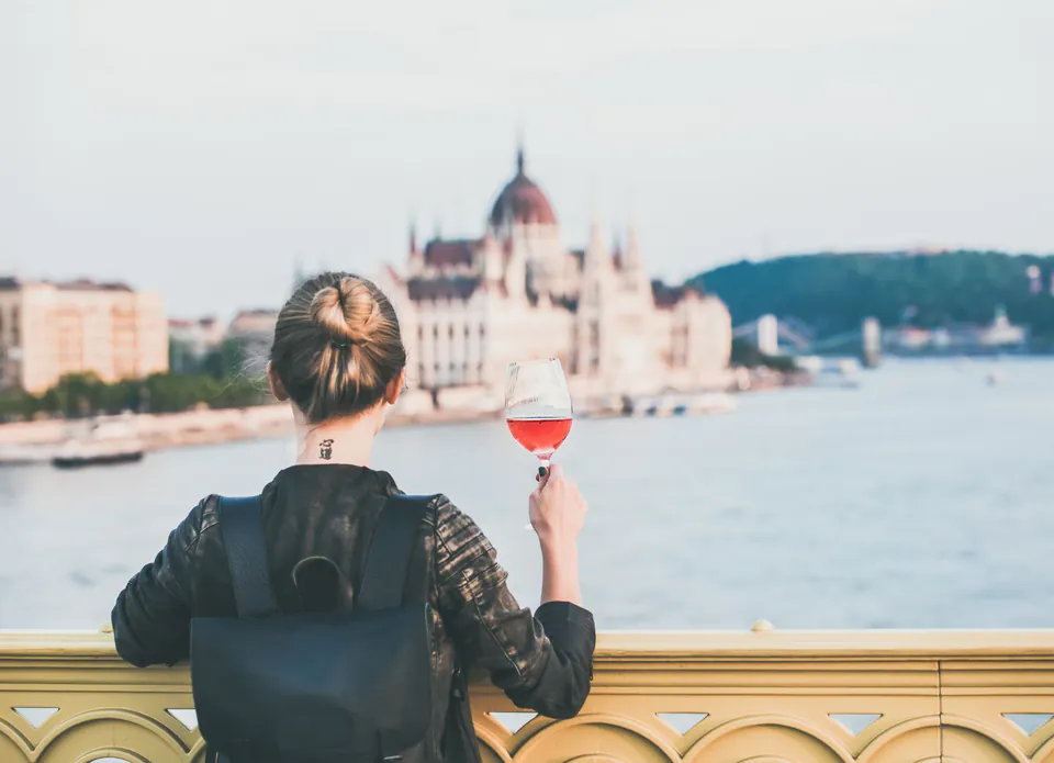Person holding wine glass overlooking Budapest Parliament building across Danube River