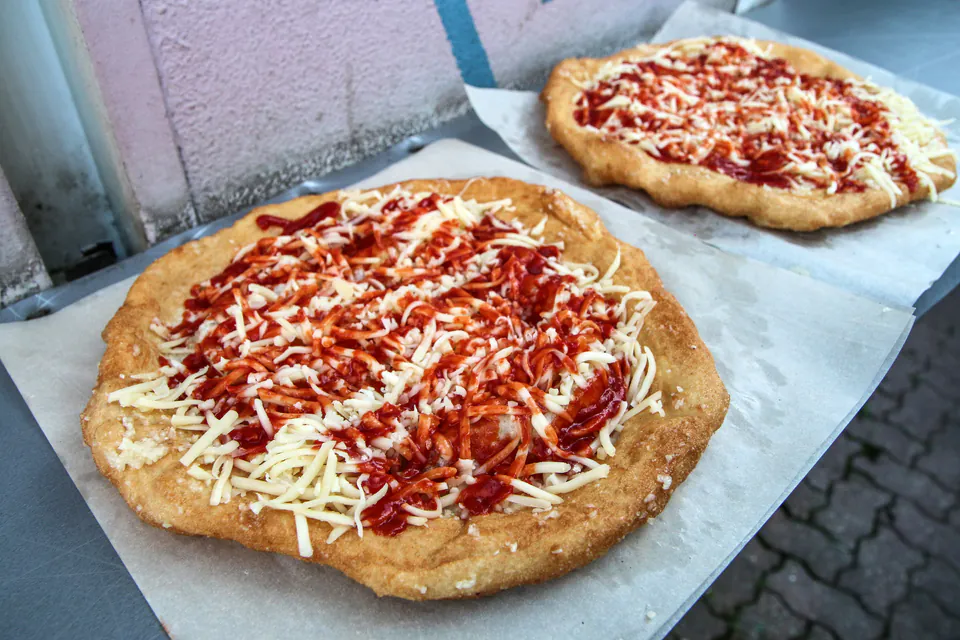 Two freshly fried lángos topped with shredded cheese and red sauce on white paper