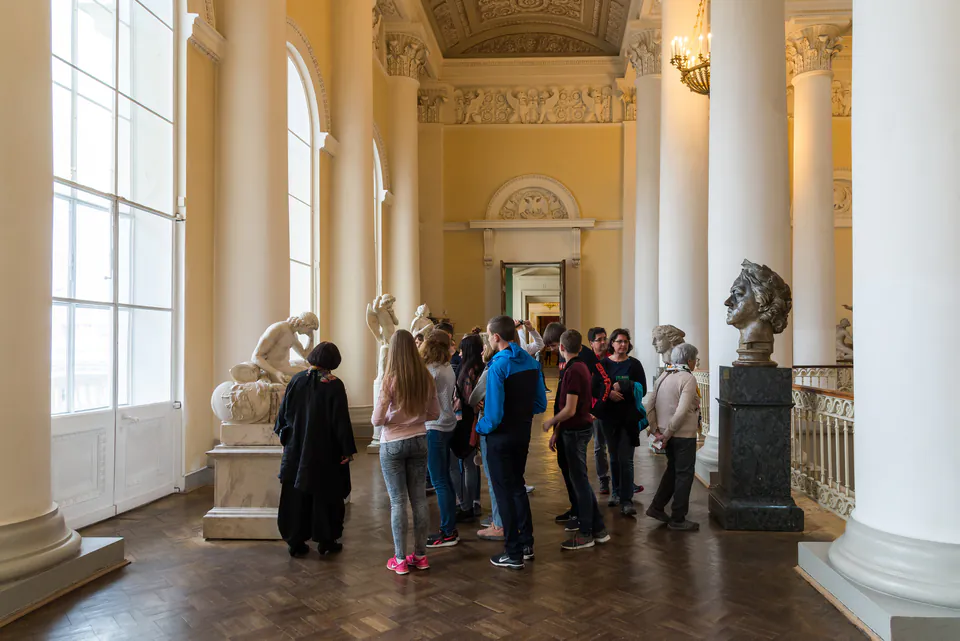 Tour group listening to guide in ornate museum hallway with classical columns and sculptures