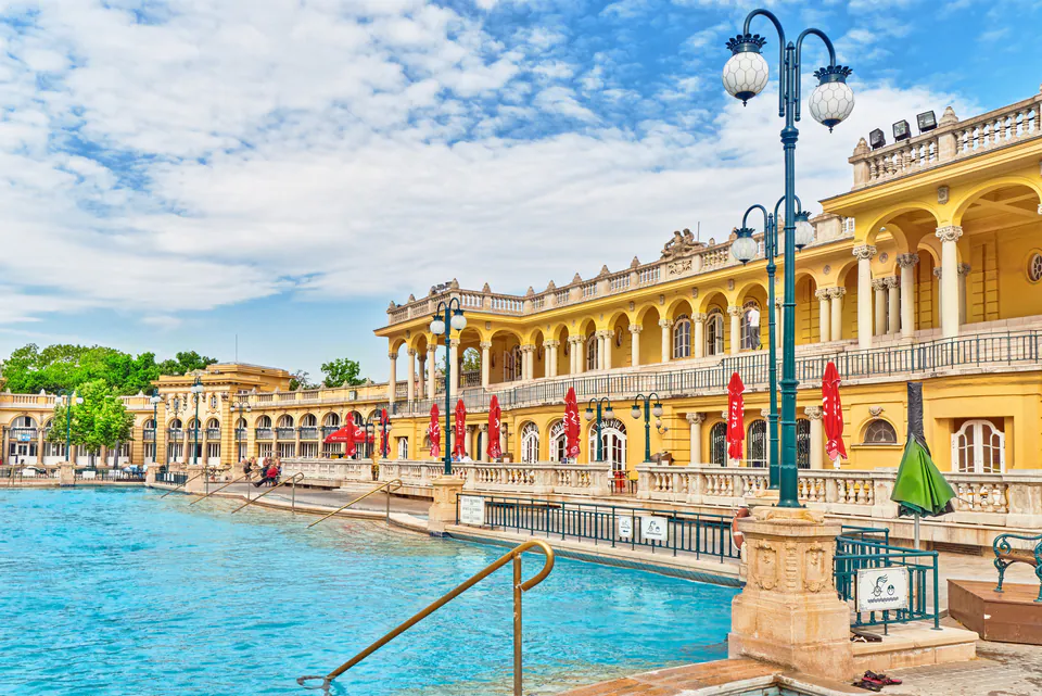 Historic yellow colonnade building alongside turquoise thermal pool with ornate lampposts