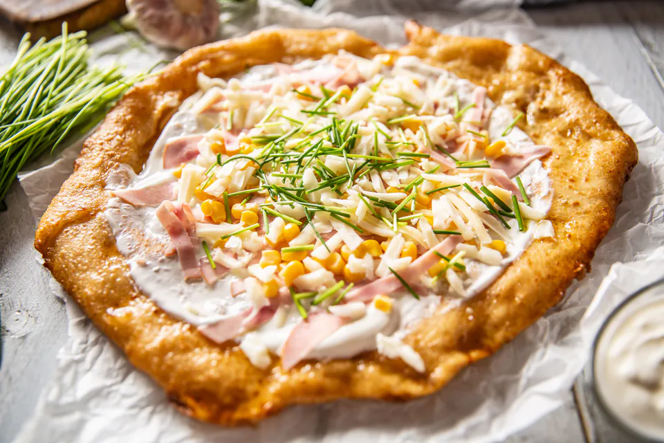 Fresh Hungarian lángos topped with sour cream, cheese, ham and chives on white paper