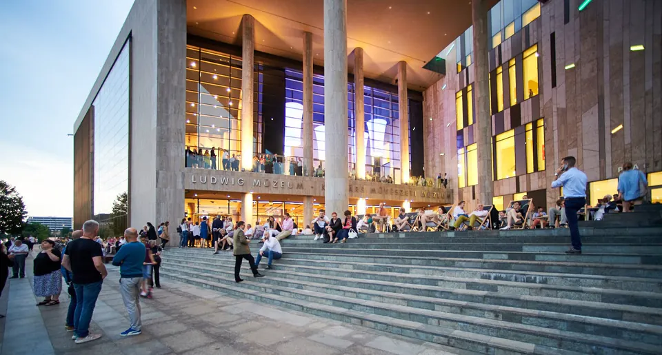 Evening crowd gathering on illuminated steps of Ludwig Museum entrance with colorful window reflections