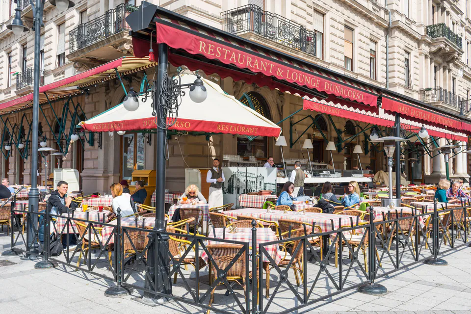 Corner restaurant with red awnings and outdoor terrace seating with checkered tablecloths