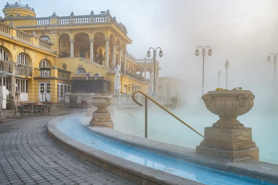 Steamy outdoor thermal pool with yellow neo-baroque building and ornate stone decorations at Széchenyi Baths