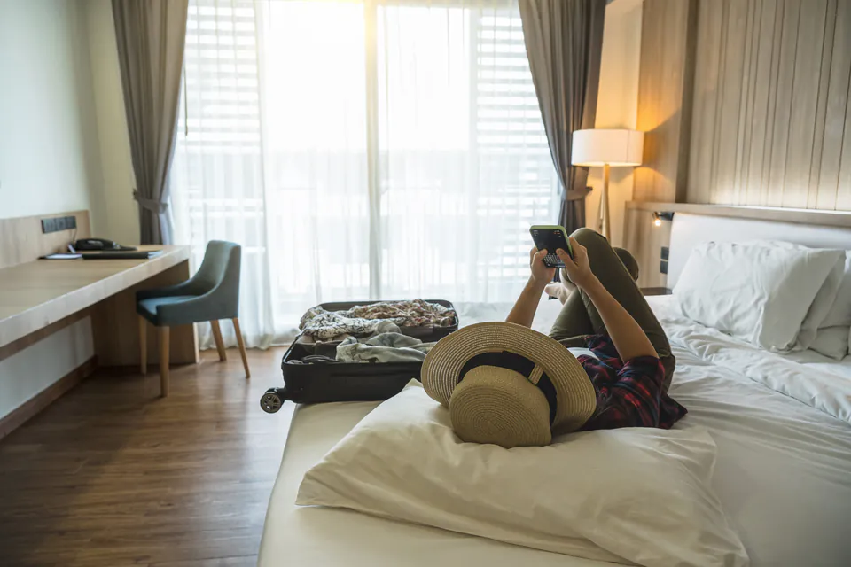 Person lying on hotel bed checking smartphone with luggage and hat nearby