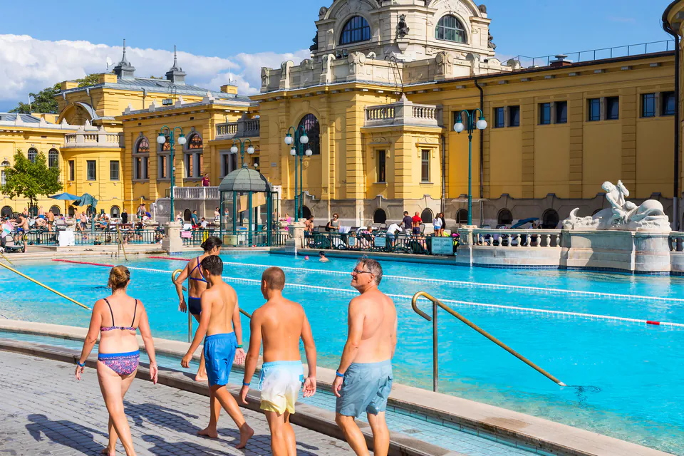 Visitors walking along the edge of Széchenyi thermal bath pool with historic yellow building in background