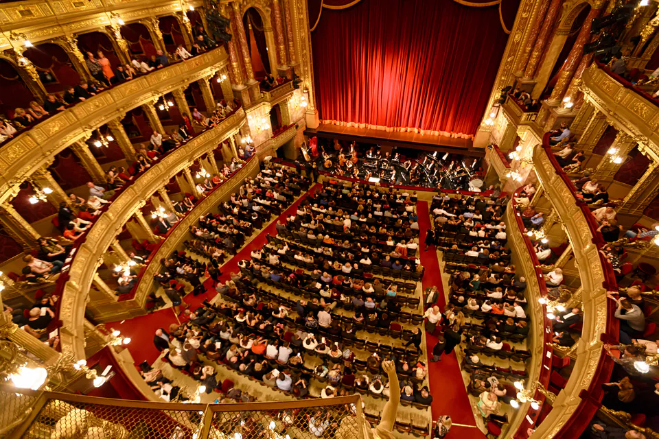 Aerial view of packed opera house auditorium with red curtain and ornate golden balconies