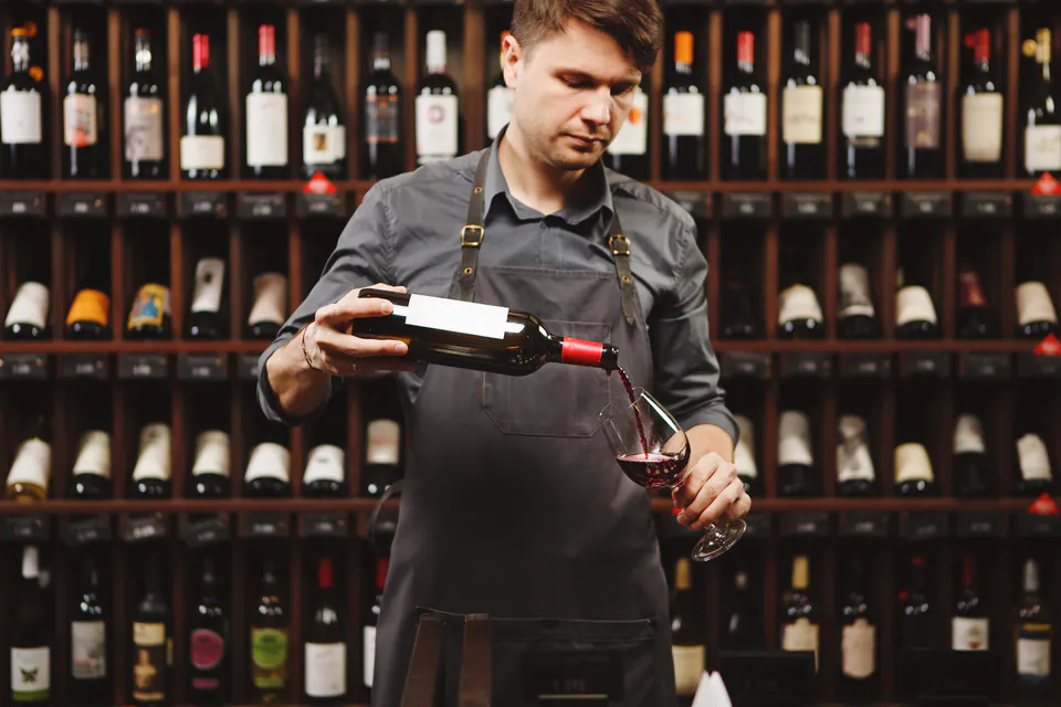 Wine professional pouring red wine into glass against backdrop of wine bottle display