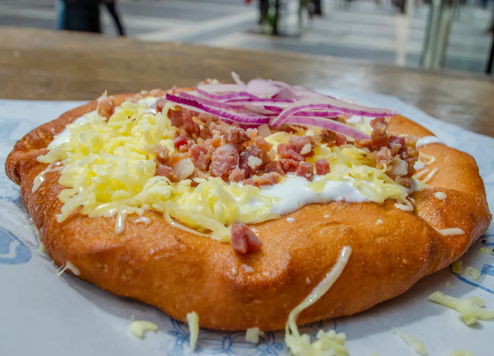 Fresh lángos topped with sour cream, shredded cheese, bacon bits and pickled red onions