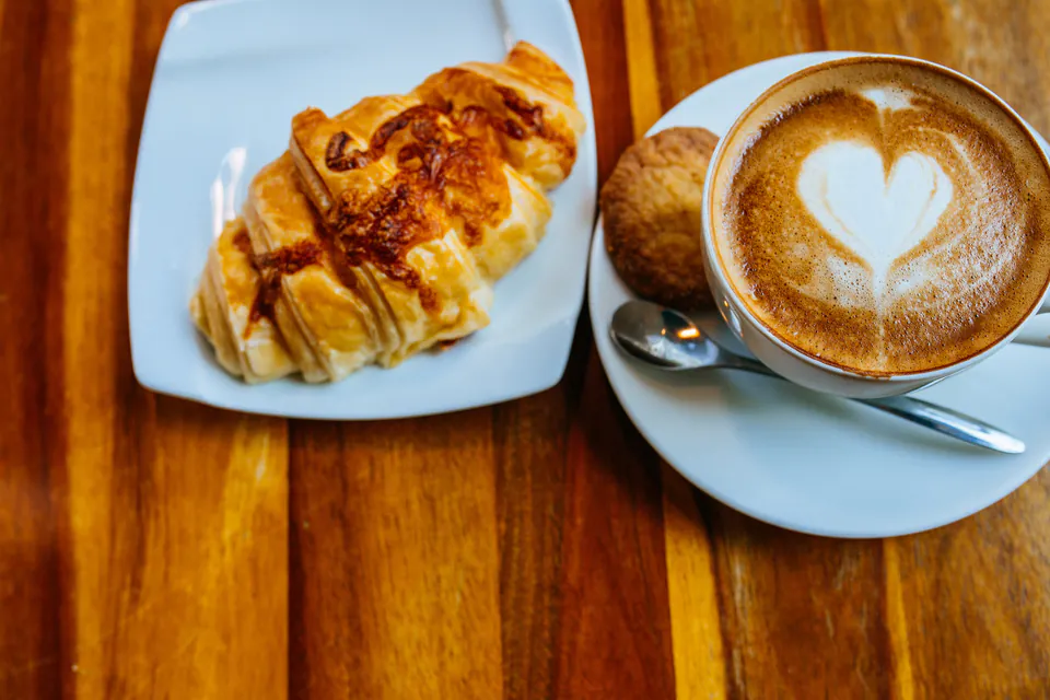 Cappuccino with heart latte art served alongside a fresh butter croissant on wooden table