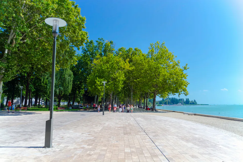 Tree-lined waterfront promenade with pedestrian walkway and lamp posts along Lake Balaton