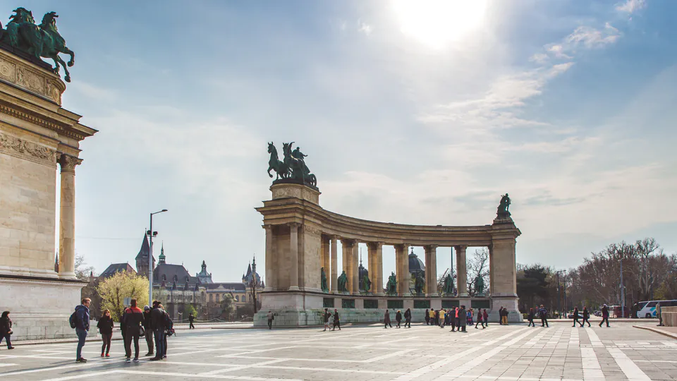 Wide view of Heroes' Square colonnade with visitors exploring the monument under sunny skies