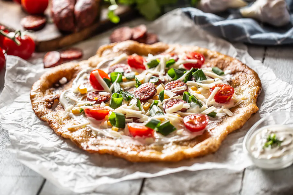 Traditional Hungarian lángos topped with sour cream, fresh vegetables and sausage slices