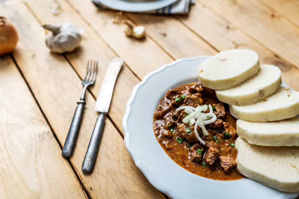Bowl of rich red goulash soup with beef chunks served alongside traditional white bread dumplings