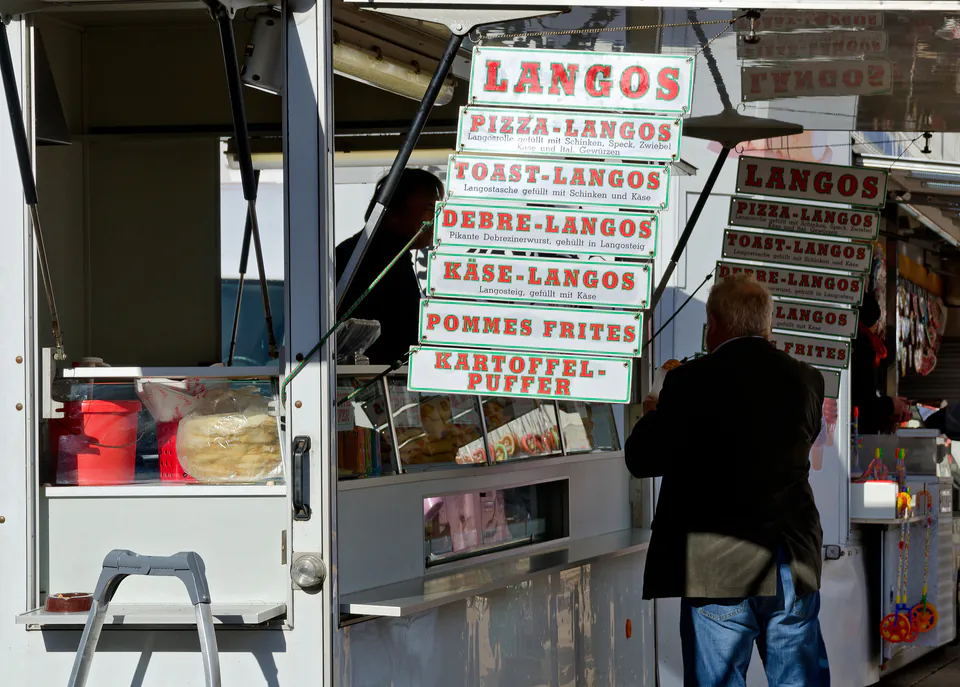 Food vendors preparing Hungarian fried bread (langos) with menu board showing toppings and prices